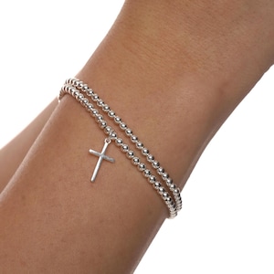 May include: A silver bracelet featuring two strands of small, round beads. A small cross pendant hangs from the bracelet. The cross is a simple, minimalist design.