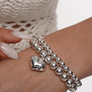 May include: A silver bracelet with a heart charm. The bracelet is made of round beads and is stacked on top of another silver bracelet.
