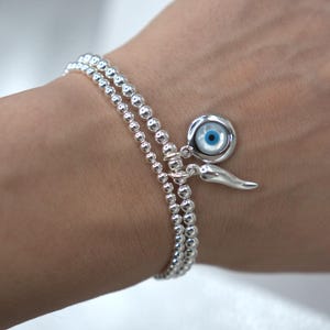 May include: A silver beaded bracelet with a charm featuring a blue evil eye and a silver horn charm.