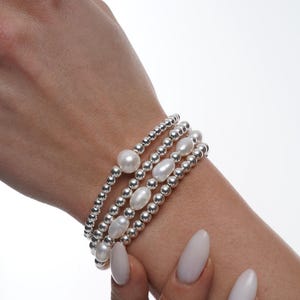 May include: A set of four silver bead bracelets with white pearl accents. The bracelets are stacked on a person's wrist.