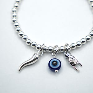 May include: Silver beaded bracelet with three charms: a silver horn, a blue and white evil eye, and a silver hand gesture. The bracelet is on a white background.