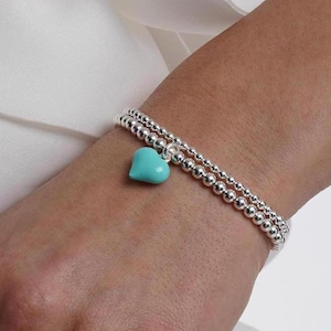 May include: Two silver bead bracelets with a turquoise heart charm on one bracelet. A silver ring with a small charm is on the index finger.