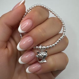 May include: Silver beaded bracelet with a cat charm. The bracelet features small, round silver beads strung together, with a detailed cat charm hanging from the center. The cat charm has a smiling face and a small bow.