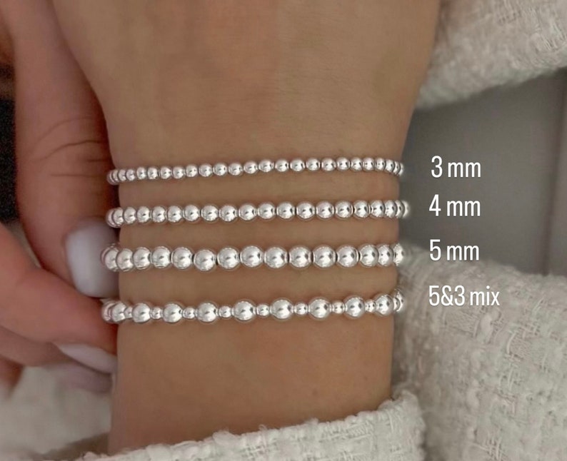 K&ouml;nnte beinhalten: Vier silberne Perlenarmb&auml;nder an einem Handgelenk. Die Armb&auml;nder bestehen aus unterschiedlich gro&szlig;en Perlen, wobei das obere Armband die kleinsten und das untere Armband die gr&ouml;&szlig;ten Perlen hat. Die Armb&auml;nder sind mit ihrer Perlengr&ouml;&szlig;e in Millimetern gekennzeichnet: 3 mm, 4 mm, 5 mm und 5 & 3 Mix.