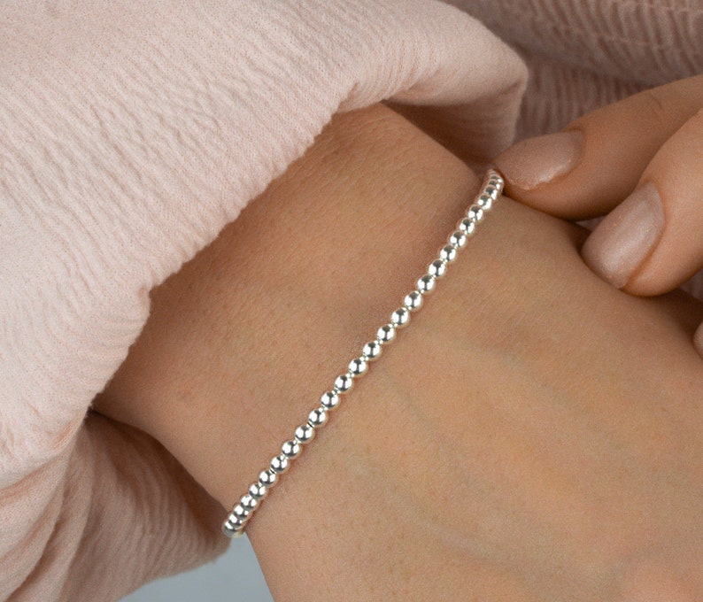 K&ouml;nnte beinhalten: Ein silbernes Perlenarmband, das an einem Handgelenk getragen wird. Das Armband besteht aus kleinen, runden Perlen, die gleichm&auml;&szlig;ig verteilt sind.