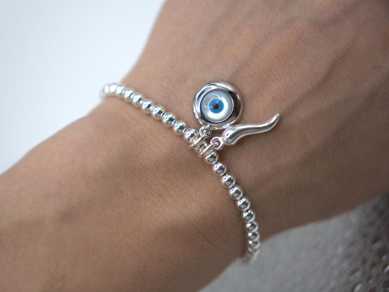 May include: A silver beaded bracelet with a charm featuring a blue evil eye and a silver horn pendant.