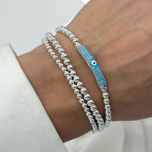 May include: Three silver beaded bracelets. One bracelet has a turquoise blue bar with a white and blue evil eye design. The bracelets are worn on a wrist, against a white shirt.