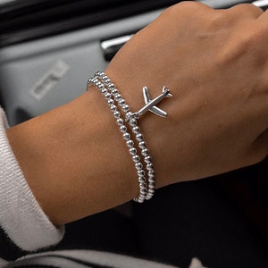 Peut inclure: Un bracelet en argent composé de perles et d'un pendentif en forme d'avion. Le bracelet est superposé et a un aspect délicat.