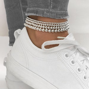 May include: A silver beaded anklet with multiple strands of varying sizes. The anklet is worn on a person's ankle.