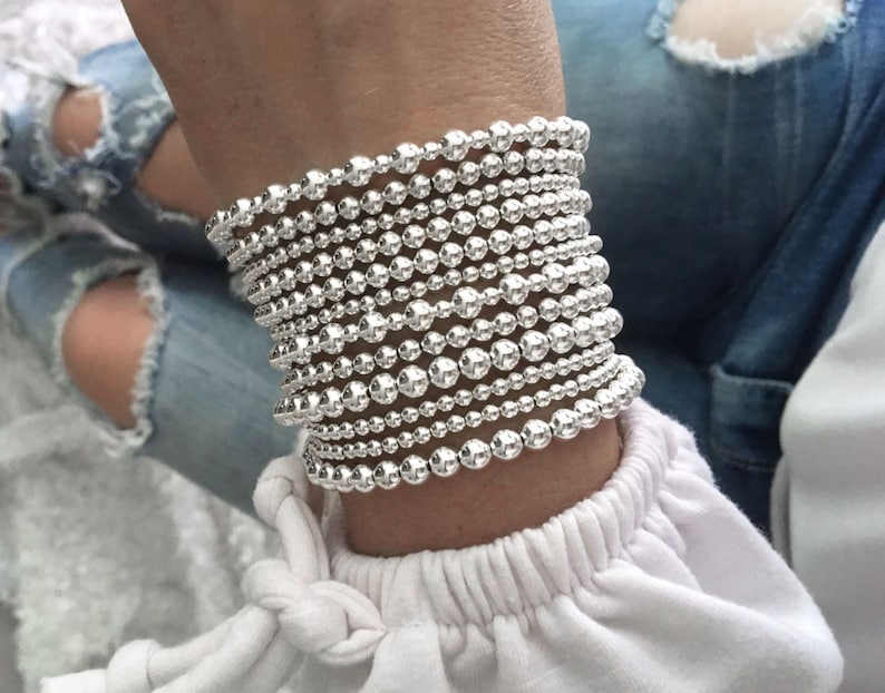 K&ouml;nnte beinhalten: Ein Stapel silberner Perlenarmb&auml;nder am Handgelenk einer Person. Die Armb&auml;nder bestehen aus kleinen, runden Perlen, die gleichm&auml;&szlig;ig verteilt sind.