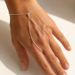 May include: A silver-toned hand chain bracelet featuring a delicate link chain that wraps around the wrist and extends to a single ring loop, connected by a small central silver bead. The design consists of thin, polished metal links.