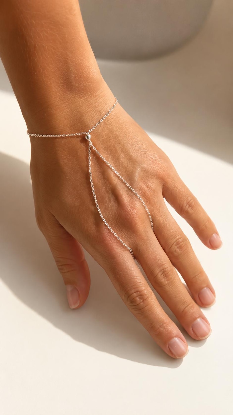 May include: A silver-toned hand chain bracelet featuring a delicate link chain that wraps around the wrist and extends to a single ring loop, connected by a small central silver bead. The design consists of thin, polished metal links.