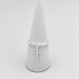 May include: A silver beaded ring with a silver cross charm. The ring is displayed on a white cone-shaped ring holder.