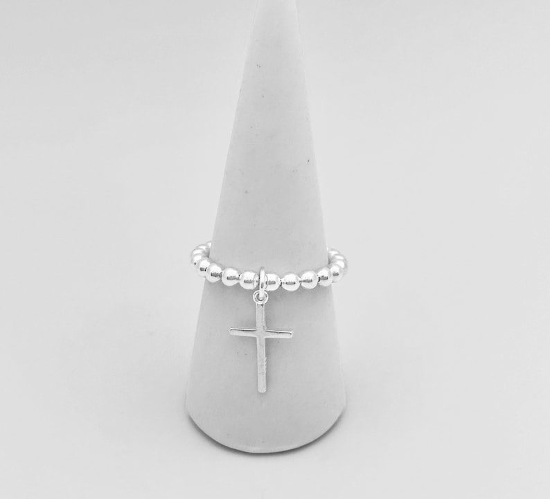 May include: A silver beaded ring with a silver cross charm. The ring is displayed on a white cone-shaped ring holder.