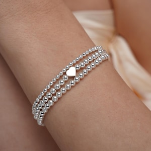 May include: A silver beaded bracelet with a heart charm. The bracelet is layered with three strands of beads.