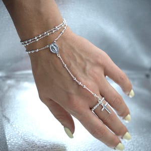 May include: A silver rosary bracelet with a ring attached by a chain. The bracelet features small, round beads and a medallion with a religious figure. The ring has a cross design.