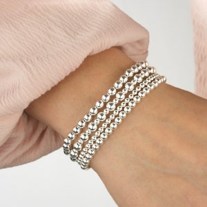 K&ouml;nnte beinhalten: Ein Set aus vier silbernen Perlenarmb&auml;ndern, die auf einem Handgelenk &uuml;bereinander liegen. Die Armb&auml;nder bestehen aus glatten, runden Perlen.