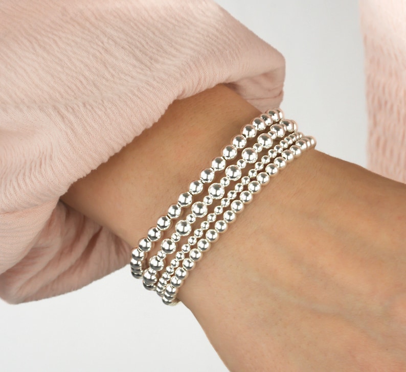 K&ouml;nnte beinhalten: Ein Set aus vier silbernen Perlenarmb&auml;ndern, die auf einem Handgelenk &uuml;bereinander liegen. Die Armb&auml;nder bestehen aus glatten, runden Perlen.