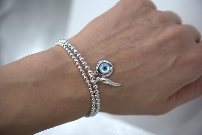 May include: A silver beaded bracelet with a charm featuring a blue evil eye and a silver horn charm.