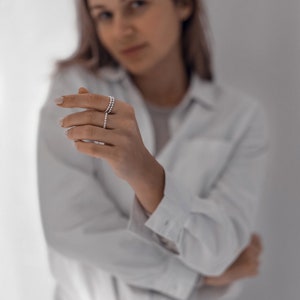 May include: A woman wearing a white button-down shirt and blue jeans is showing off a silver ring on her finger.