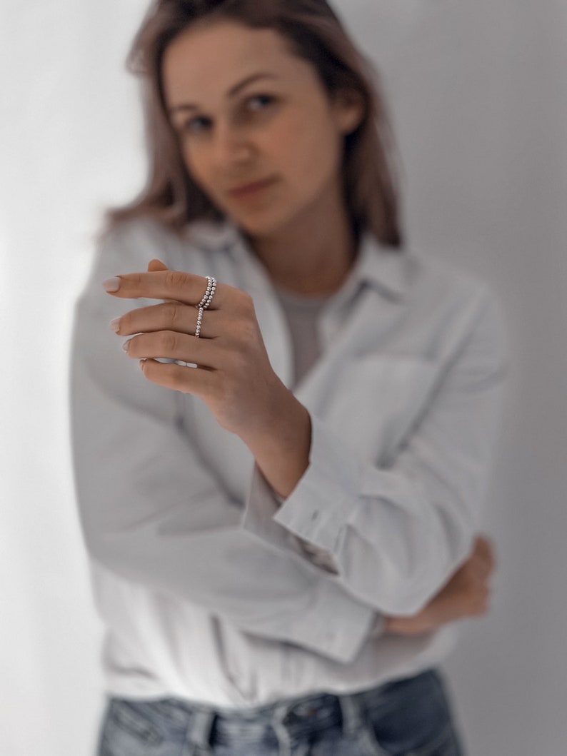 May include: A woman wearing a white button-down shirt and blue jeans is showing off a silver ring on her finger.