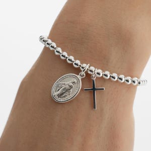 May include: A silver beaded bracelet featuring two charms: an oval pendant with a religious figure and a small cross. The bracelet is displayed on a wrist.