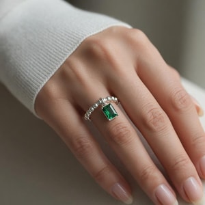 May include: A silver ring featuring a beaded band and a single emerald-cut green gemstone charm dangling from the centre. The ring is crafted with a polished metallic finish, showcasing a vibrant green rectangular stone set in a simple silver frame.