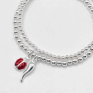 May include: Two silver beaded bracelets with charms. One bracelet features a red ladybird charm, and the other has a silver horn charm. The bracelets are on a white background.