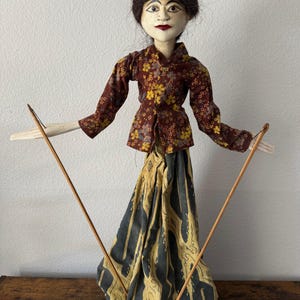 May include: A traditional Indonesian Wayang Golek puppet. The doll has a painted face, dark hair, and is dressed in a maroon floral top and a patterned skirt. The puppet is held with wooden rods.