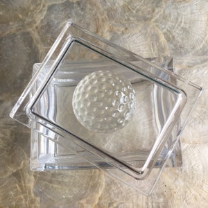 Op de afbeelding: Een transparante glazen doos met een golfbal erin. De doos heeft een deksel dat lichtjes openstaat.