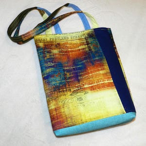 May include: A rectangular tote bag with a colorful abstract design. The bag features a mix of yellow, orange, blue, and brown hues, with the words "GREAT PORTLAND STREET" visible. It has blue accents and handles.