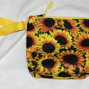 May include: A small, square zippered pouch with a vibrant sunflower pattern. The pouch has a yellow zipper and a matching yellow strap. The sunflowers are yellow and orange with dark brown centers.