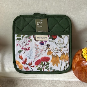 Puede incluir: Una manopla de cocina cuadrada de color verde oliva con textura acolchada y un bolsillo frontal blanco con un estampado botánico de flores rojas, amarillas, rosas y helechos. Una etiqueta dice OVEN MITT 1 PACK 20X20CM y SHAXU GIFT A LITTLE BIT OF JOY.