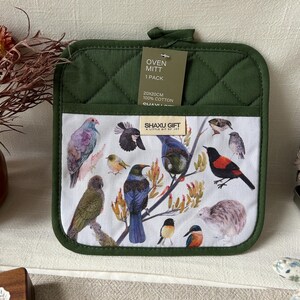 May include: A square, dark green quilted oven mitt featuring a white pocket printed with various colorful bird illustrations. A tag on the mitt reads OVEN MITT 1 PACK 20X20CM 100% COTTON, and a small label displays the text SHA XU GIFT A LITTLE BIT OF JOY.