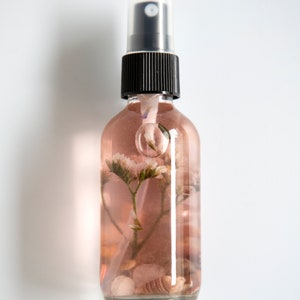 May include: A clear glass bottle with a black spray top filled with a pink liquid and dried flowers and seashells.