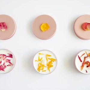 May include: Six small round candles with dried flower petals embedded in the wax. Three candles have pink petals and three candles have yellow petals. The candles are in individual containers with pink lids.