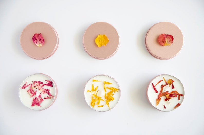 May include: Six small round candles with dried flower petals embedded in the wax. Three candles have pink petals and three candles have yellow petals. The candles are in individual containers with pink lids.