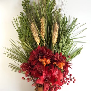 May include: A bouquet of dried flowers and greenery, including red dahlias, red alstroemeria, wheat stalks, and sprigs of greenery. The bouquet is tied together with cinnamon sticks and twine.