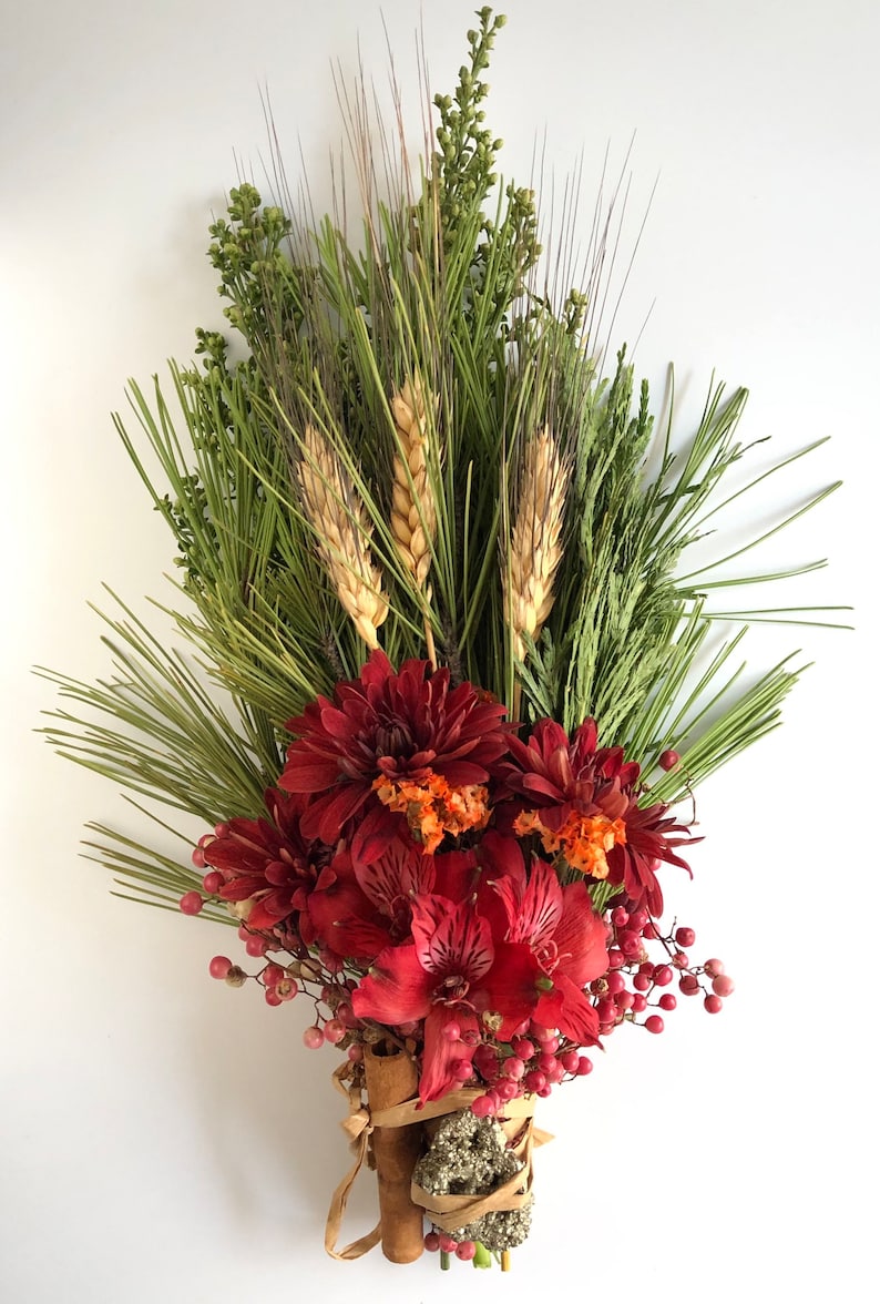 May include: A bouquet of dried flowers and greenery, including red dahlias, red alstroemeria, wheat stalks, and sprigs of greenery. The bouquet is tied together with cinnamon sticks and twine.