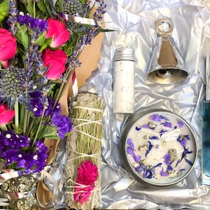 Puede incluir: Una caja de regalo llena de un ramo de flores rosas y moradas, una vela blanca con detalles morados y dorados, una pequeña botella de sales de baño blancas, una campana plateada y una botella de vidrio azul con tapa plateada.