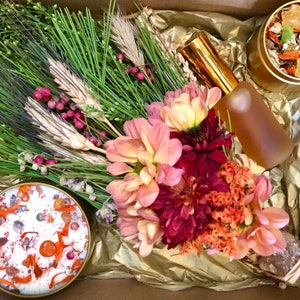 Puede incluir: Una caja de regalo llena de un ramo de flores secas, una vela con detalles naranja y blanco, una botella de líquido ámbar y una lata de hierbas secas y cristales.