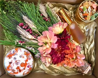 May include: A gift box filled with a bouquet of dried flowers, a candle with orange and white accents, a bottle of amber liquid, and a tin of dried herbs and crystals.