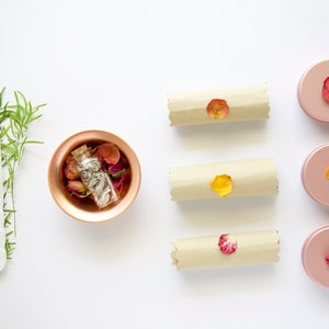 May include: A flat lay of a copper bowl filled with dried rose petals and a small glass bottle, three rolled up pieces of parchment paper with dried rose petals on top, and four pink round containers with dried rose petals on top.