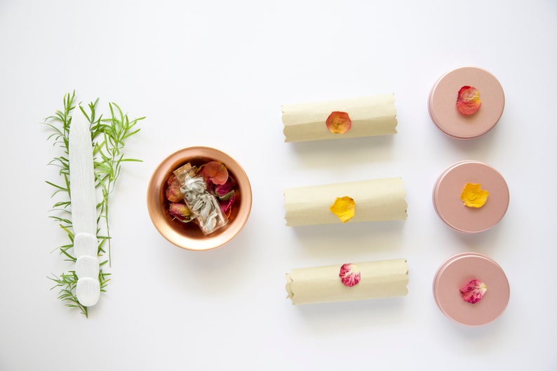 May include: A flat lay of a copper bowl filled with dried rose petals and a small glass bottle, three rolled up pieces of parchment paper with dried rose petals on top, and four pink round containers with dried rose petals on top.