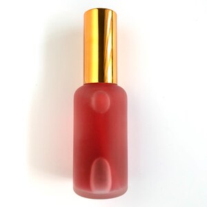 May include: A frosted glass bottle with a gold cap, filled with a pink liquid. The bottle is standing upright on a white surface.