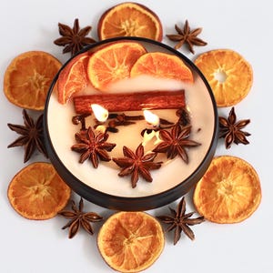 Puede incluir: Una vela encendida en un cuenco negro, rodeada de rodajas de naranja secas y anís estrellado. La vela tiene una ramita de canela y rodajas de naranja encima. Un centro de mesa cálido y acogedor.