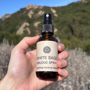 Pu&ograve; includere: Un flacone spray in vetro marrone con erogatore nero ed etichetta beige. L'etichetta presenta un'icona mandala circolare, il testo "WHITE SAGE SMUDGE SPRAY" e le parole "cleansing~clarifying~healing" stampate in inchiostro nero.