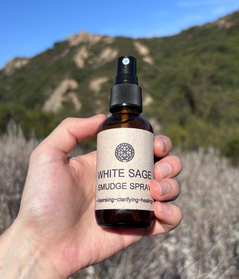 Pu&ograve; includere: Un flacone spray in vetro marrone con erogatore nero ed etichetta beige. L'etichetta presenta un'icona mandala circolare, il testo "WHITE SAGE SMUDGE SPRAY" e le parole "cleansing~clarifying~healing" stampate in inchiostro nero.