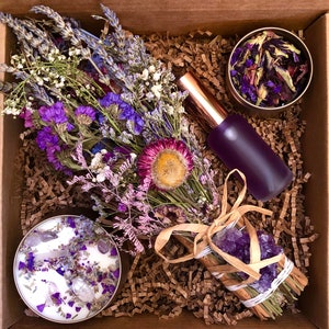 Puede incluir: Una caja de regalo llena de flores secas moradas y lavanda, una vela morada con cristales, una pequeña lata de flores secas y una botella de líquido morado.