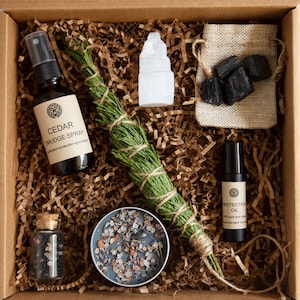 Può includere: Una scatola regalo piena di oggetti per un rituale di protezione. La scatola contiene uno spray di incenso di cedro, un bastoncino di incenso, un cristallo trasparente, una piccola borsa di pietre nere, una bottiglia di olio protettivo e un piccolo barattolo di cristalli neri.